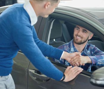 Man smiling in car dealership shaking hands with a salesperson through the driver’s window, illustrating a rent‑to‑own car agreement option for people without Good Credit.