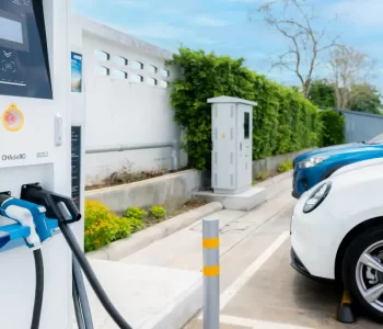 Electric vehicle charging station with a white EV plugged in beside parked cars, highlighting EV infrastructure contrasted with ICE vehicles in everyday Australian driving context.