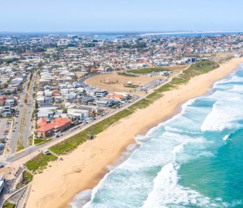 Aerial view of a coastal city with a sandy beach, ocean waves, and roads along the shoreline under a clear sky—capturing Newcastle, where CarCoop expands its Rent-to-Own Car Service to Newcastle & Wollongong.