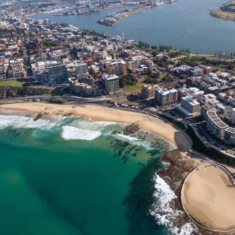 Aerial view of a coastal city with sandy beaches, ocean waves, waterfront buildings, and a river or harbor in the background—perfect for exploring in Bad Credit Rent-to-Own Cars in Newcastle.