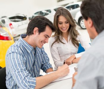 Couple signing paperwork with a car dealer while choosing a vehicle through a rent to own car scheme in a showroom.